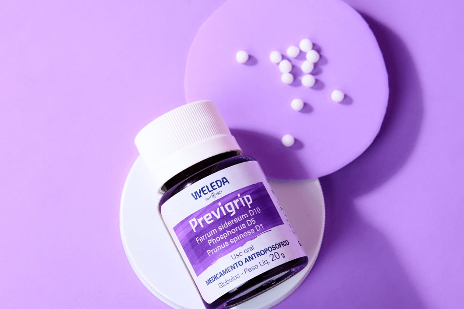 Frasco do medicamento antroposófico Previgrip da marca Weleda sobre fundo lilás, com glóbulos brancos espalhados ao redor.