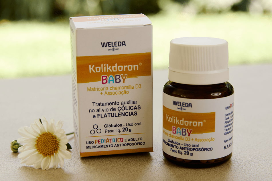 Medicamento Kolikdoron Baby da Weleda com embalagem e frasco expostos sobre superfície clara, ao lado de flor de camomila, indicado para alívio de cólicas e flatulências em bebês e adultos.