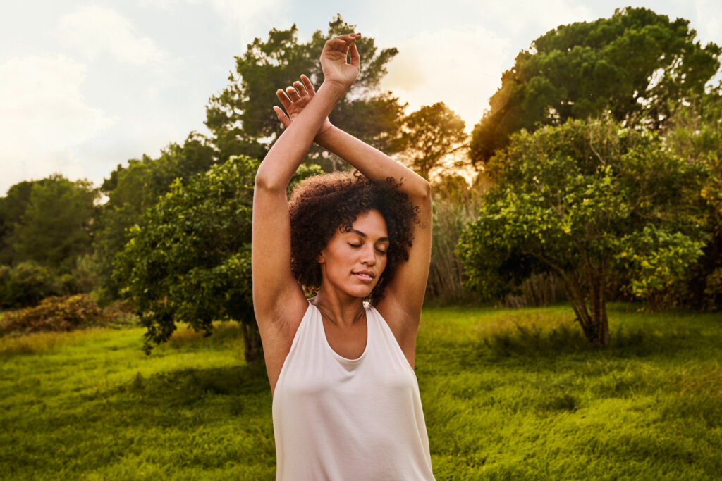 Mulher em pé ao ar livre, com os braços erguidos e olhos fechados, praticando alongamento em meio a um campo verde com árvores e luz do sol ao fundo.