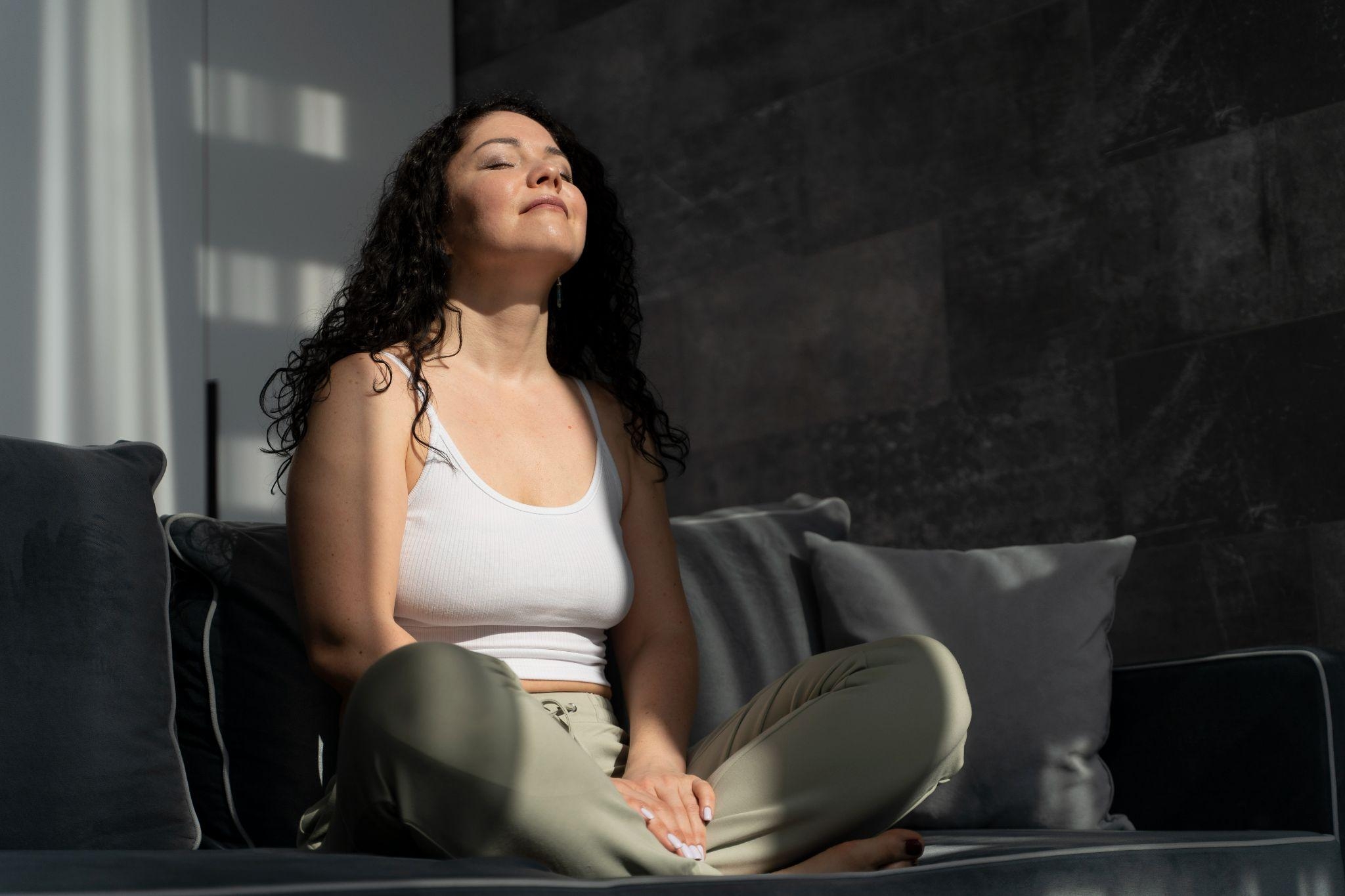 Mulher sentada em posição de meditação no sofá, com as pernas cruzadas, usando regata branca e calça clara, em sala iluminada pela luz natural que entra pela janela.